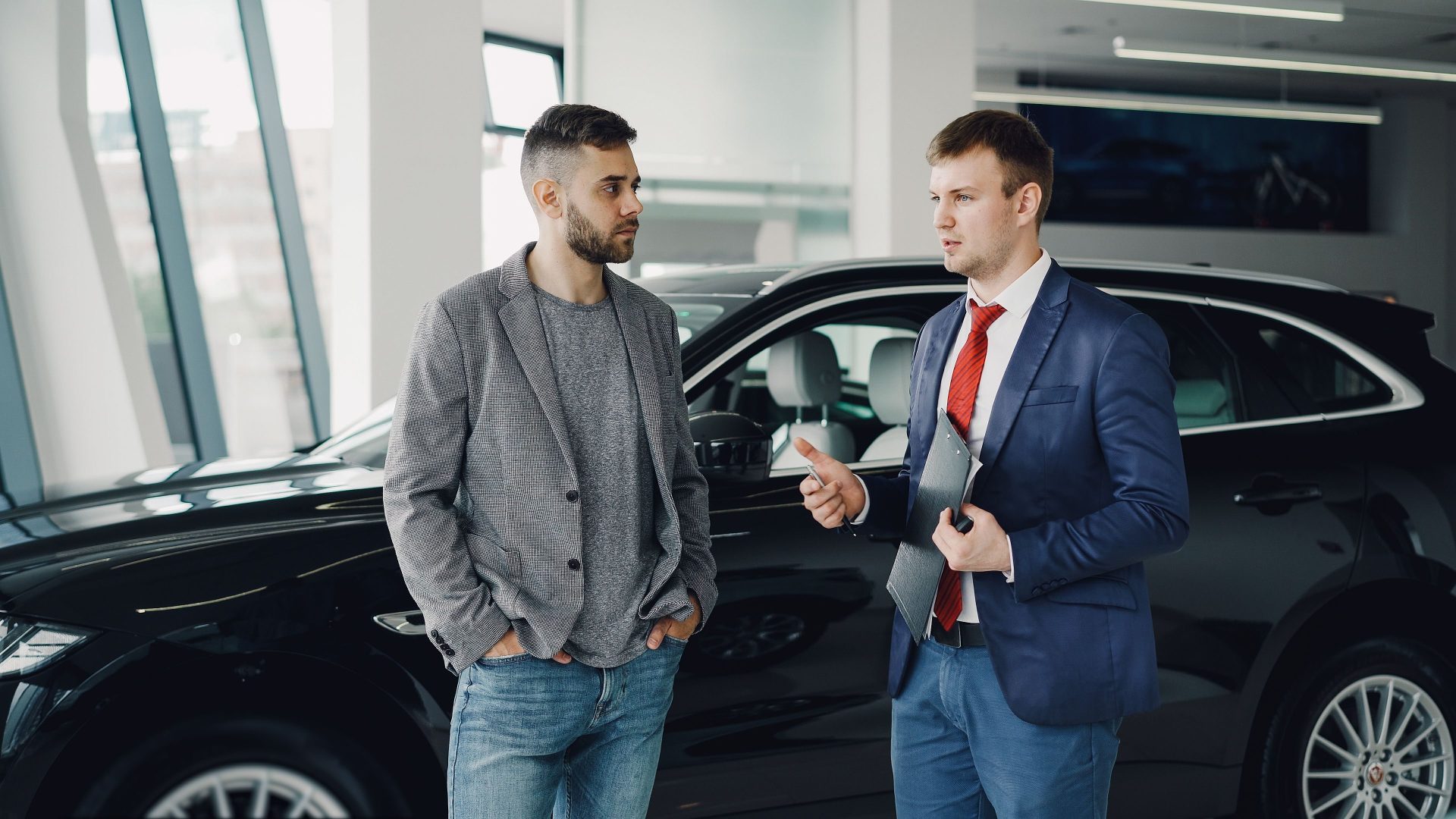 Zwei Männer unterhalten sich in einem Autohaus über Geschäfte und stehen neben einem Fahrzeug.